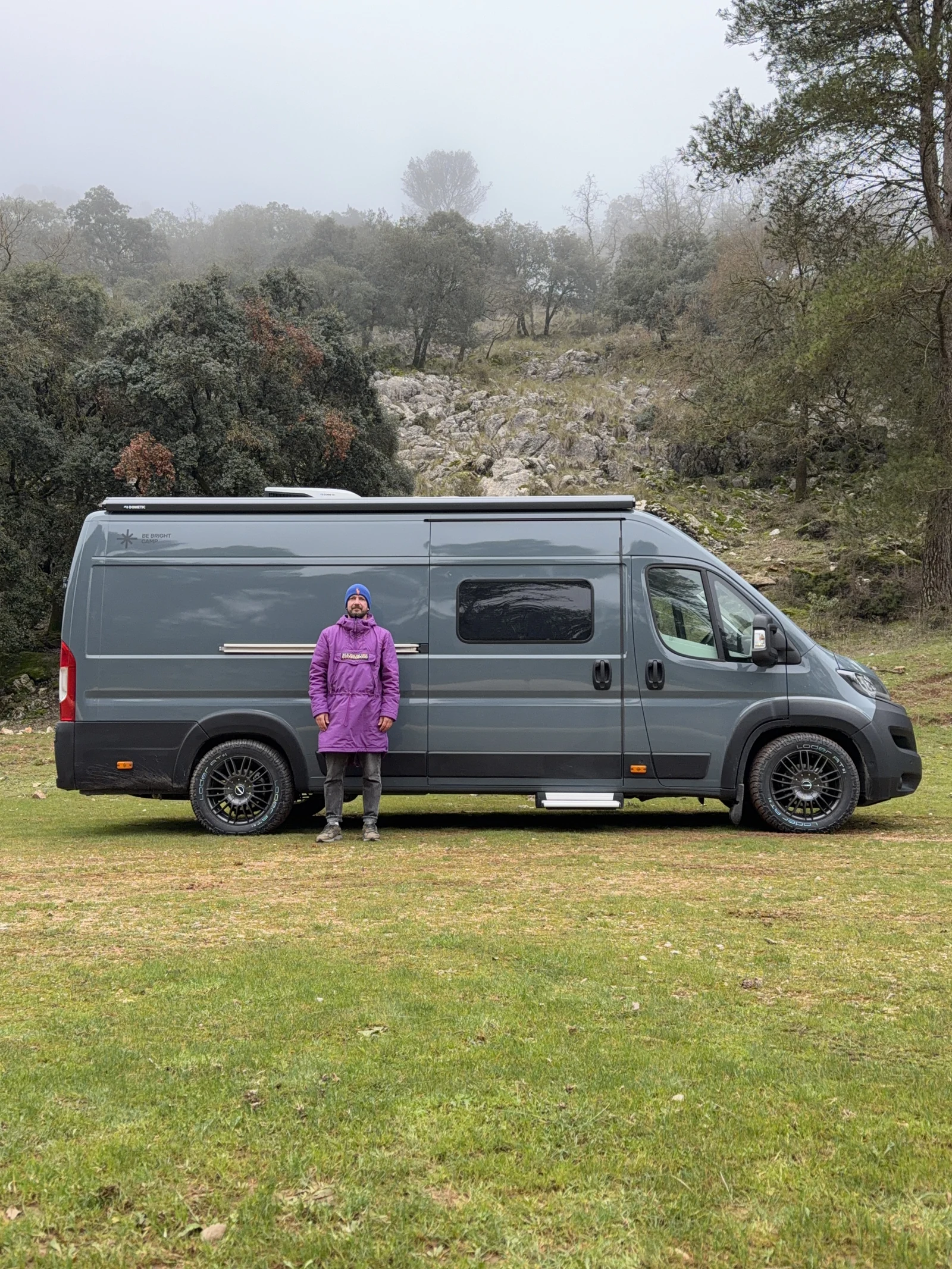 Traveller beside a grey BeBrightCamp campervan in a misty wooded landscape